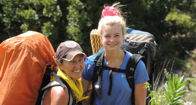 Two people wearing backpacks smile for the photo in front of green trees. 